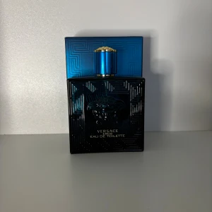 Versace Eros Eau de Toilette sampel - Versace Eros Eau de Toilette i en lyxig blå glasflaska med guldiga detaljer och Medusa-huvudet ingraverat på framsidan. Flaskan har en fyrkantig form och ett elegant mönster som ger en exklusiv känsla. Perfekt för dig som gillar stilrena och ikoniska dofter.