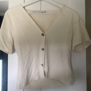 Beige kortärmad blus från 157 - Stilren beige blus från 157 med korta ärmar och v-ringning. Toppen har unika silvriga knappar framtill och är lätt croppad. Perfekt för dig som gillar minimalistisk och clean stil. Mjuk och skön bomullskänsla.
