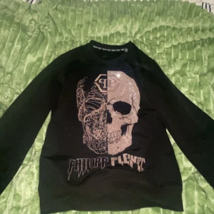 Svart Philipp Plein tröja med döskalle - Svart långärmad tröja från Philipp Plein med en stor glittrig döskalle framtill och logga under. Tröjan har rund halsringning och är i mjukt material, perfekt för dig som gillar edgy och unik stil. Äkta!