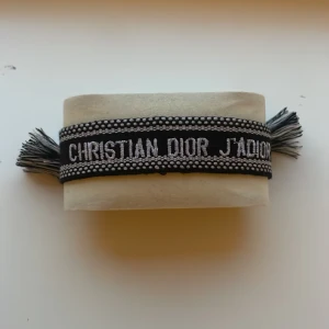 Christian Dior svart tygarmband - Snyggt svart tygarmband från Christian Dior med broderad text 'CHRISTIAN DIOR J'ADIOR' i vitt. Armbandet har vita prickdetaljer längs kanterna och fransar i ändarna för en cool touch. Perfekt statement piece för dig som gillar lyxiga accessoarer.