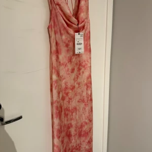 Rosa långklänning - Säljer en helt oanvänd långklänning i rosa satin. Klänningen har draperad halsringning, öppen rygg med knytband och är ärmlös. Prislappen finns kvar. 