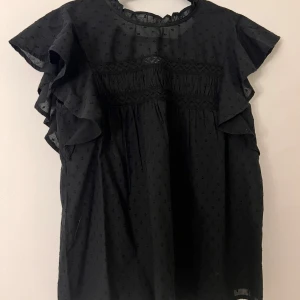 Svart blus med volang från Vero Moda - Supersöt svart blus från Vero Moda med volangärmar och diskret prickigt mönster. Blusen har rund halsringning, spetsdetaljer och är luftig i modellen. 