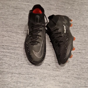 Nike phantom GT2 elite FG  - Säljer ett par Nike Phantom GT fotbollsskor i svart med orange detaljer på dobbarna och röd insida. Skorna har snörning, syntetmaterial och en modern design med vit Nike-logga på sidan. Perfekta för dig som vill dominera på planen.