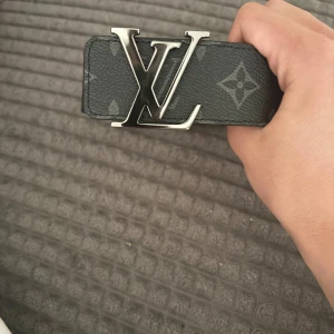 LV bälte med box och kvitto - Säljer ett svart Louis Vuitton-bälte i skinn med klassiskt monogrammönster och stor silvrig LV-logga som spänne. Bältet har en bred design och är tillverkat i Spanien. Perfekt för dig som vill ha en statement-accessoar. UTAN BOX 1500 eller 1K vid snabb affär