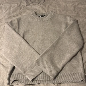 Grå stickad tröja från Gina Tricot - En stilren grå stickad tröja från Gina Tricot med rund halsringning och ribbade muddar vid ärmslut och nederkant. Perfekt för lager-på-lager och enkel att matcha med jeans eller kjol. Klassisk passform och mjukt material.