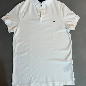 Vit Tommy Hilfiger piké - Klassisk vit pikétröja från Tommy Hilfiger med korta ärmar och liten broderad logga på bröstet. Tröjan har krage och knäppning med tre knappar. Tillverkad i mjuk bomull som känns skön mot huden. Perfekt för en clean och stilren look.