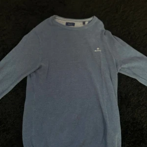 Blå stickad tröja från GANT - Snygg blå stickad tröja från GANT med diskret logga på bröstet. Tröjan har rund halsringning, ribbade muddar och är långärmad. Perfekt för lager-på-lager och passar till jeans eller chinos.