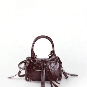 Vinröd handväska med dragkedja - Snygg vinröd handväska i glansigt skinn med silverfärgade metalldetaljer och dragkedja framtill. Väskan har dubbla handtag, avtagbar axelrem och coola långa läderband som dekoration. Perfekt storlek för mobilen och andra småsaker.