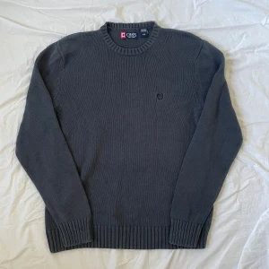 Mörkgrå stickad tröja från Chaps - Mörkgrå stickad tröja från Chaps i 100% bomull. Klassisk modell med rund halsringning, ribbade muddar och diskret broderad logga på bröstet. Perfekt för lager-på-lager och chill stil.