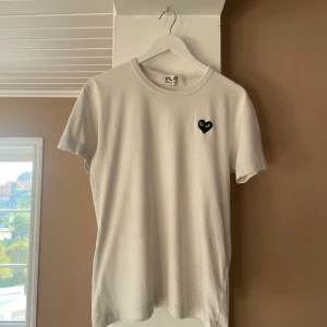Vit t-shirt Comme des Garçons Play - Vit t-shirt från Comme des Garçons Play med klassisk svart hjärtlogga på bröstet. Enkel och stilren design med rund hals och korta ärmar. Tillverkad i mjuk bomull som känns skön mot huden. Perfekt för dig som gillar minimalistisk streetstyle. Den är i storlek M men passar S. Tröjan är i mycket fint skick!