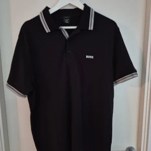 Svart pikétröja från BOSS - Svart pikétröja från BOSS med vit logga på bröstet. Klassisk krage och knäppning framtill, samt vita och grå ränder på krage och ärmslut. Tillverkad i bomull för en skön känsla. Perfekt för en clean och stilren look.