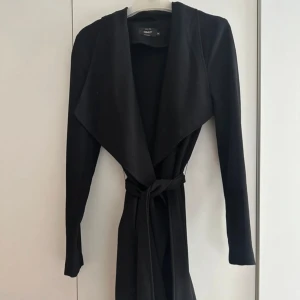 Svart kappa från ONLY med bälte - Svart kappa från ONLY med krage och knytskärp i midjan. Jackan har lång ärm, är ofodrad och är tillverkad i ett mjukt tyg som ger en clean och stilren look. Perfekt för dig som gillar minimalistisk stil och vill ha en jacka som sticker ut på ett chill sätt