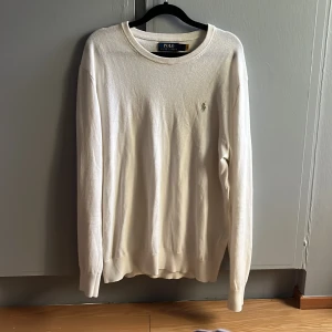 Vit stickad tröja från Polo Ralph Lauren - Klassisk vit stickad tröja från Polo Ralph Lauren med rund halsringning och broderad logga på bröstet. Tröjan har lång ärm och ribbade muddar, perfekt för en clean och stilren look. Den är i nyskick!