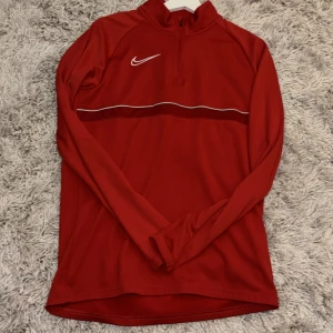 Röd Nike Dri-Fit half zip träningströja - Nike Dri-Fit långärmad träningströja i rött med vit broderad swoosh på bröstet och en tunn vit linje över bröstet. Tröjan har half zip och är gjord i ett lätt, syntetiskt material som andas. Perfekt för sport eller löpning. Hör av er om ni vill ha fler bilder! Pris kan diskuteras.
