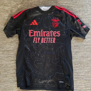 Svart Benfica fotbollströja Adidas - Snygg svart Benfica fotbollströja från Adidas med röda detaljer och tryck. Tröjan har korta ärmar, mönster av stjärnbilder och loggor från Emirates och Sagres. Tillverkad i lätt och ventilerande material med AEROREADY-teknologi. ❗️ÄKTA❗️