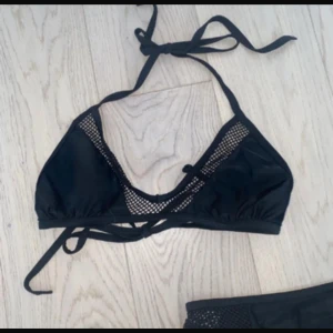 Svart bikini med meshdetaljer - Snygg svart bikini med triangelformad topp och meshdetaljer längs kanterna. Toppen har knyt både i nacken och runt ryggen för justerbar passform. Materialet är mjukt och följsamt, perfekt för strandhäng eller poolparty. Stilren och modern design som verkligen sticker ut.