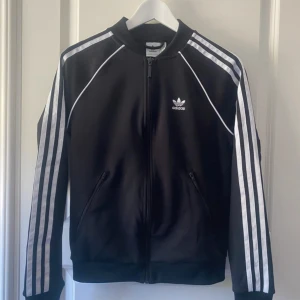 Svart Adidas bomberjacka med vita ränder - Svart Adidas bomberjacka med klassiska vita ränder längs ärmarna och vit logga på bröstet. Jackan har dragkedja framtill, ribbade muddar och är tillverkad i polyester. Perfekt för en sportig och stilren look.