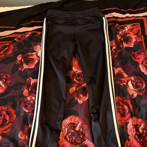 Svarta Adidas mjukisbyxor med vita ränder - Svarta mjukisbyxor från Adidas med klassiska vita ränder längs sidorna. Byxorna har resår i midjan och är gjorda i ett mjukt, syntetiskt material som känns skönt mot huden. 