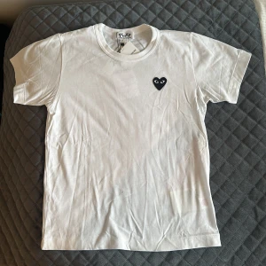 Vit t-shirt Comme des Garçons Play - Vit t-shirt från Comme des Garçons Play med klassiskt svart hjärta med ögon broderat på bröstet. T-shirten är i mjuk bomull och har en enkel, clean look med rund hals och korta ärmar. Perfekt för dig som gillar stilrena streetwear-plagg.