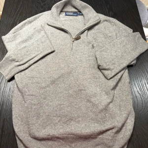 Grå halvziptröja från Polo Ralph Lauren - Snygg grå stickad tröja från Polo Ralph Lauren med halv dragkedja och broderad logga på bröstet. Tröjan har hög krage och ribbade muddar. Perfekt för lager-på-lager och chill stil.