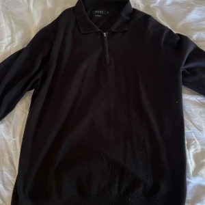 Svart tröja - Svart stickad polotröja från Riley med krage och half zip-dragkedja framtill. Tröjan har lång ärm och klassisk passform, perfekt för lager-på-lager under kyliga dagar.