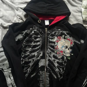 Ed Hardy hoodie med skelett och rosor - Svart hoodie från Ed Hardy med grått skelett-tryck över hela tröjan, dödskalle och röda rosor på framsidan. Röd insida i huvan och dragkedja framtill. Ikonisk Ed Hardy-logga på ryggen. Perfekt för dig som gillar streetwear med attityd.