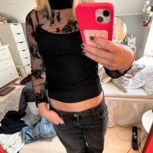 Svart mesh blus med spetsdetaljer - Säljer en svart mesh blus med långärmade transparenta ärmar och blommiga spetsdetaljer. Toppen har en tight passform och hög hals, perfekt för att ge en edgy och snygg look. Materialet är lätt och stretchigt, vilket gör den bekväm att bära.