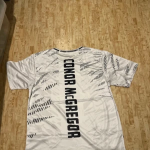 Vit UFC Conor McGregor t-shirt Venum - Säljer en vit UFC t-shirt från Venum med Conor McGregor-tryck på ryggen och coola svarta grafiska detaljer. Framsidan har UFC-logga, Venum-logga och crypto.com-tryck. T-shirten är kortärmad och har sportig vibe, perfekt för dig som gillar fighting och streetstyle.
