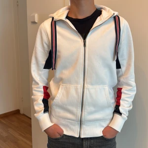 Vit hoodie Tommy Hilfiger S - Vit hoodie från Tommy Hilfiger med dragkedja, huva och snygga röda och marinblå detaljer på ärmarna. Stora broderade loggan på ryggen och randiga snören i huvan. Mjuk bomullskänsla och två fickor framtill. Perfekt för en sportig och stilren look.