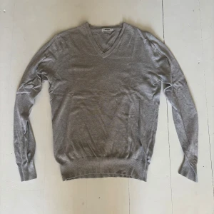 Grå v-ringad tröja J.Lindeberg - Grå v-ringad tröja från J.Lindeberg med diskret logga på bröstet. Tröjan är i mjuk bomullsmix och har långa ärmar samt ribbade muddar vid ärmslut och nederkant. Perfekt för en clean och stilren look.