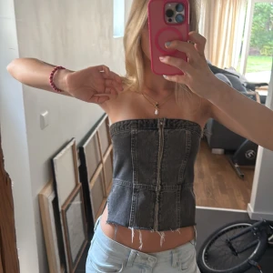 Svart jeans bandeau tubtopp - Säljer en svart bandeau tubtopp i jeansmaterial med rå kant nertill och dragkedja framtill. Toppen är croppad och har en cool, edgy vibe som passar perfekt till baggy jeans eller kjol.
