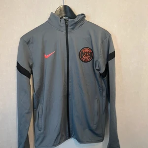 Grå PSG träningsjacka från Nike - Säljer en grå träningsjacka från Nike med Paris Saint-Germain-logga i orange och svart på bröstet. Jackan har hel dragkedja, lång ärm och svarta detaljer på ärmarna. Perfekt för dig som gillar fotboll och vill ha en sportig look.