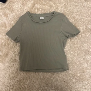 Ribbad grön t-shirt från Lindex - Snygg ribbad t-shirt från Lindex i en dov grön färg. Modellen är kortärmad med rund hals och har en skön, stretchig passform. Perfekt att matcha med jeans eller shorts för en chill look.