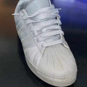 Adidas Superstar i helvitt skinn - Klassiska Adidas Superstar sneakers i helvitt skinn med ikonisk snäcktå och tre ränder på sidan. Skorna har platt sula, snörning och en stilren look som passar till allt. Perfekta för dig som gillar tidlös streetstyle.