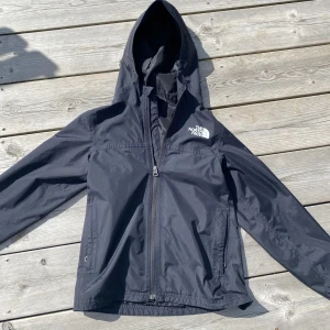 Svart vindjacka från The North Face - Svart vindjacka från The North Face med huva och hel dragkedja framtill. Jackan har två sidofickor med dragkedja och en vit logga på bröstet. Tillverkad i lätt och slitstark polyester, perfekt för blåsiga dagar. Original pris 899kr
