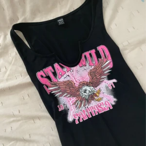 Svart linne med örntryck från SHEIN - Coolt svart linne från SHEIN med tryck av en örn och texten 'STAY WILD' i rosa och vitt. Linnet har djup rundad halsringning och ärmlös design. Materialet är mjukt och stretchigt, perfekt för en edgy look med streetkänsla.