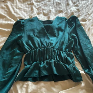 Grön omlottblus med volang - Snygg grön omlottblus med långa puffärmar och markerad midja med rynk och volangkant nedtill. Blusen har v-ringning och är tillverkad i ett glansigt, mjukt material som ger en lyxig känsla. Perfekt för dig som vill sticka ut med en cool och trendig look.