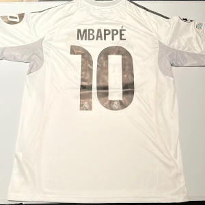 Real Madrid Mbappé 10 matchtröja 2024 - Säljer en officiell Real Madrid fotbollströja från Adidas med Mbappé och nummer 10 på ryggen. Tröjan är vit med svarta detaljer, guldiga inslag och klubbmärke på bröstet. Champions League-märke på ärmen och 'Emirates Fly Better' tryck på framsidan. Materialet är lätt och andas. Replika, endast handtvätt gäller!