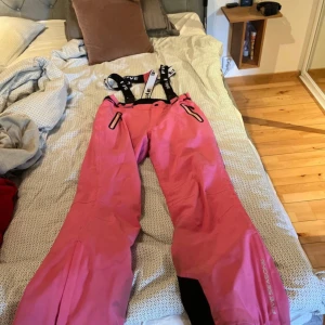Rosa skidbyxor   från Five Seasons - Rosa skidbyxor från Five Seasons med svarta och vita hängslen. Byxorna har dragkedjor vid fickorna och förstärkning vid bensluten. Perfekta för vintern och har en snygg sportig look. Materialet är syntet och byxorna är fodrade för extra värme. Slitna längst ner men funkar lika bra ändå. 