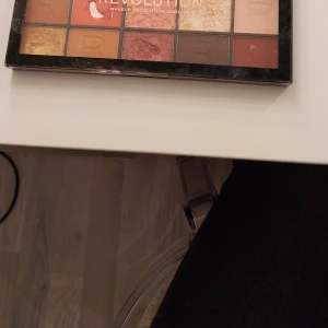 Revolution Reloaded Smokey Desire Palett - Säljer en Revolution Reloaded Smokey Desire ögonskuggspalett med 15 pressade puder-skuggor i varma och neutrala toner. Färgerna inkluderar guld, koppar, röd, rosa, beige och bruna nyanser. Perfekt för att skapa både mjuka och intensiva looks. Paletten är kompakt och har genomskinligt lock.