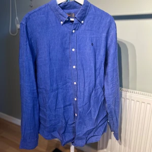 Blå linneskjorta från Morris  - Snygg blå skjorta i linne från Morris  med klassisk button-down krage och vita knappar. Skjortan har lång ärm och en liten broderad logga på bröstet. Perfekt för dig som gillar en stilren och avslappnad look.