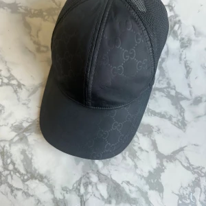 Svart Gucci keps med GG-mönster - Snygg svart keps från Gucci med diskret GG-mönster på framsidan och mesh på baksidan. Justerbar snapback för perfekt passform. Märkt med Gucci-logga både invändigt och bak. Perfekt för dig som vill ha en stilren och lyxig accessoar.
