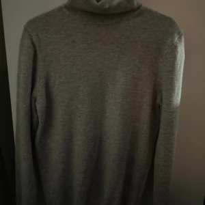 Grå polotröja i kashmir från Massimo Dutti - Supermjuk och stilren grå polotröja från Massimo Dutti i 100% kashmir. Klassisk modell med lång ärm och ribbade muddar. Perfekt för lager-på-lager och ger en clean vibe till din outfit.