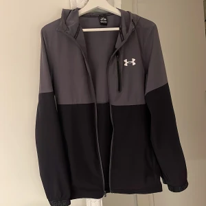 Under Armour vindjacka  - Säljer en Under Armour vindjacka i mycket bra skick! Denna jacka är perfekt för alla typer av utomhusaktiviteter, oavsett om det är löpning, vandring eller bara en promenad i stan.   Färg: Svart och grå Skick: Mycket bra, nästan som ny Fickor: 3 rymliga fickor med plats för telefon, nycklar och andra värdesaker  Jackan är både lätt och slitstark, vilket gör den idealisk för varierande väderförhållanden.