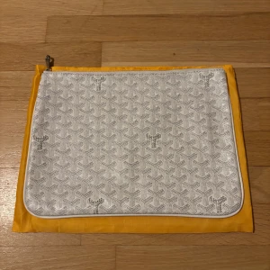 Vit Goy4rd handväska - Säljer en stilren vit handväska med deras ikoniska mönster och silverfärgad dragkedja. Väskan är i skinn och har en clean look med Goy4rd-logga tryckt på flera ställen. Kommer med original gul dustbag. Helt ny. Om du har tur så kan du få plats med en liten bärbar dator. Går att använda som necessär väska.