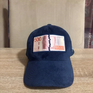 Blå keps Bruce Lee x All Cap One - Snygg mörkblå keps med mesh på sidorna och broderad text. Framtill sitter en patch med motiv av en 500-kronorssedel. Justerbar baktill för perfekt passform. Perfekt statement piece för dig som gillar unika accessoarer.