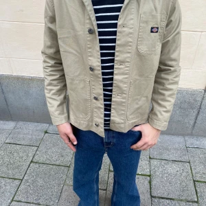 Beige overshirt från Dickies - Säljer en beige overshirt från Dickies med klassisk knappstängning och tre praktiska fickor framtill. Jackan har en stilren look med lång ärm och diskret logga på bröstfickan. Perfekt för dig som gillar enkel och clean streetstil.
