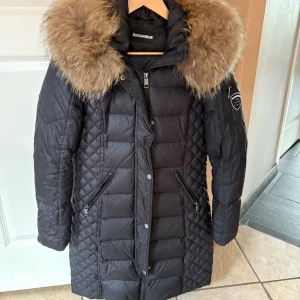 Svart dunjacka Rockandblue päls - Svart lång dunjacka från Rockandblue med stor fluffig fuskpäls runt huvan. Jackan har quiltad design, dragkedjor på fickor och ärmslut samt patch på ena ärmen. 