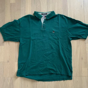 Grön pikétröja från Tommy Hilfiger - Snygg grön pikétröja från Tommy Hilfiger med klassisk krage och knappslå. Tröjan har korta ärmar och ett broderat emblem på bröstet. Materialet är mjuk bomull som känns skönt mot huden. Perfekt för dig som gillar stilrena och tidlösa plagg.