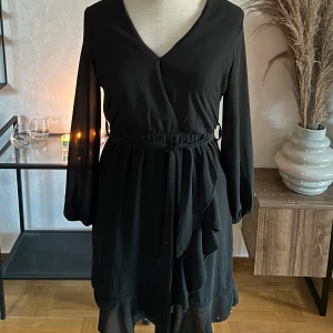 Svart omlottklänning med volang - Stilren svart omlottklänning med v-ringning, långa transparenta ärmar och knytband i midjan. Kjolen har volangdetalj och lätt flowy känsla. Perfekt för dig som gillar en klassisk look med lite extra edge.